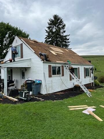 Roofers working on a white house with brown shutters and a damaged roof, on a cloudy day.