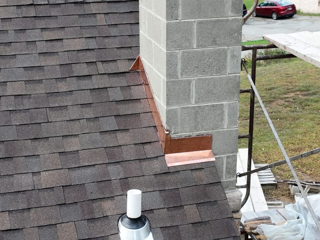 Copper flashing on a brown shingle roof next to a gray cinder block chimney.