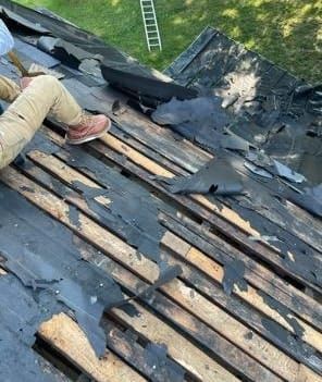 Person on a roof removing old shingles, revealing wooden planks. Green grass and ladder in the background.