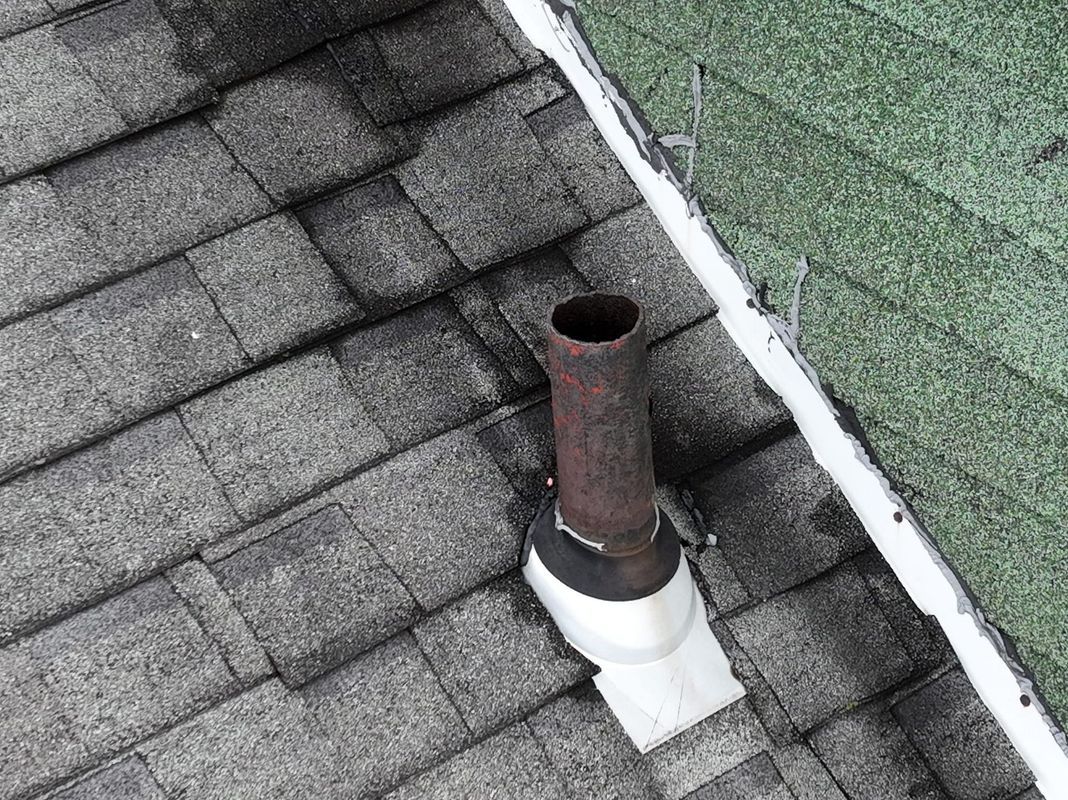 Rusty pipe on a gray shingle roof, adjacent to a green shingle roof with white trim.
