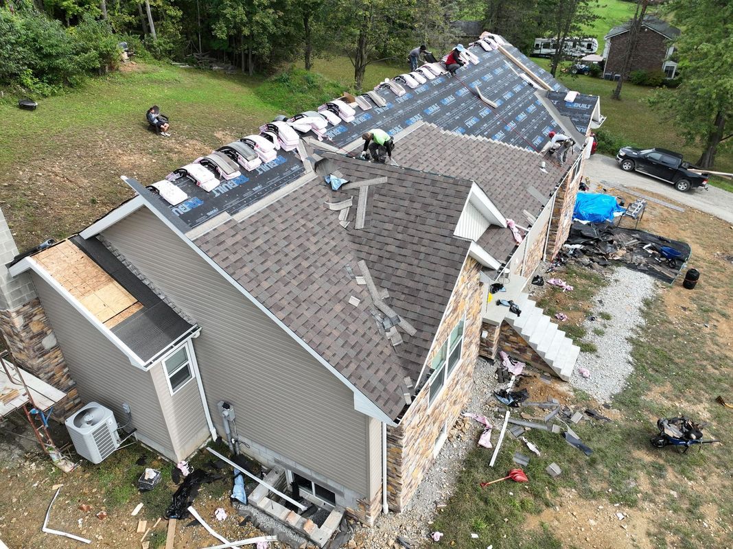 Roofers replacing shingles on a house with tan siding and a stone facade, near a driveway.