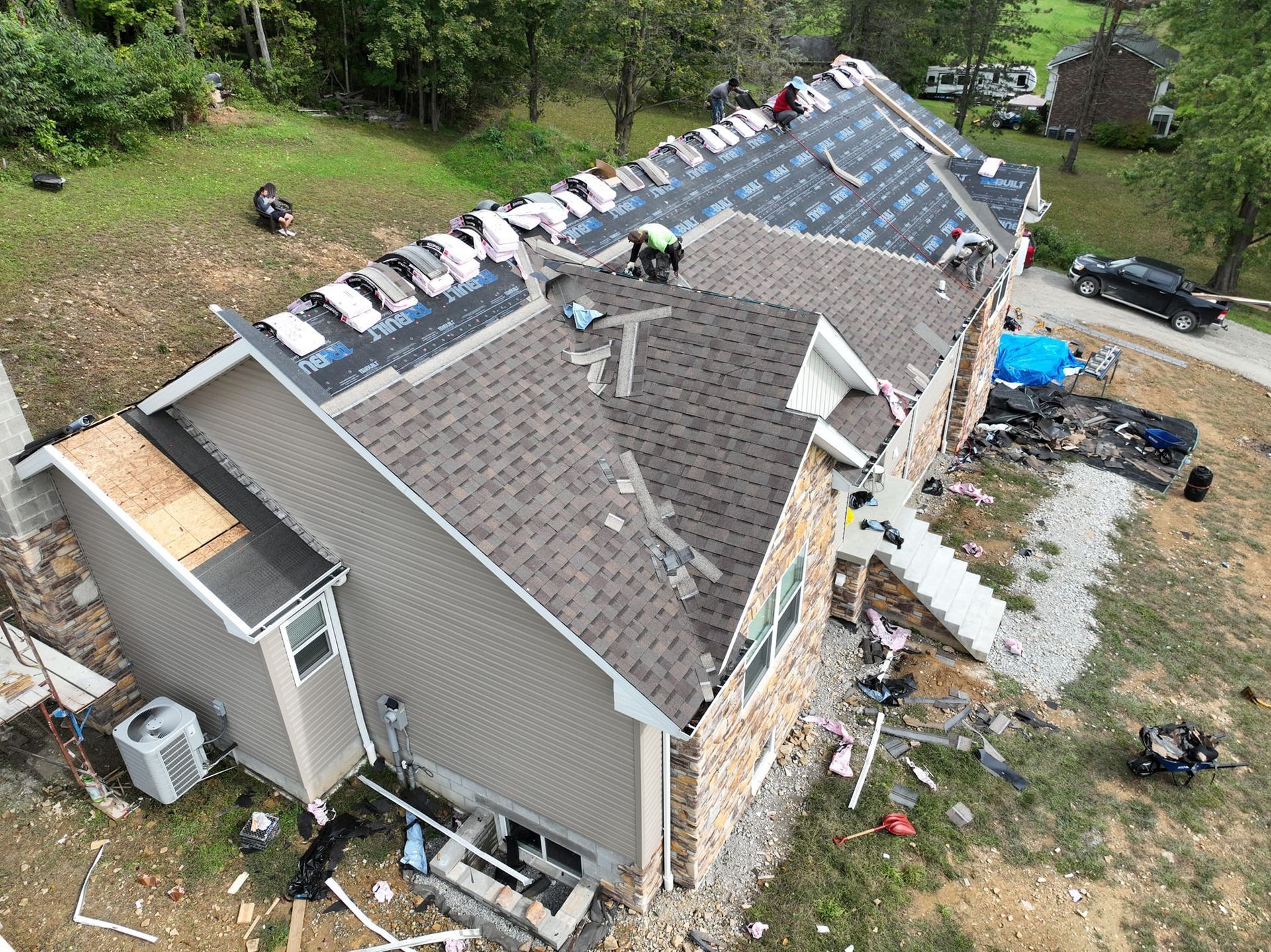 Roofers working on a house roof, laying down shingles. Green trees and parked vehicles surround the home.