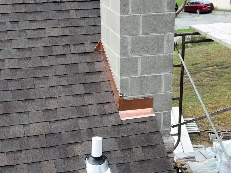 Roof with asphalt shingles meeting a concrete chimney. Copper flashing present. Scaffolding in background.