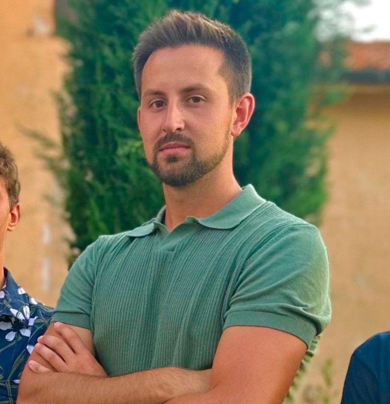 Man with crossed arms, wearing a green polo shirt, standing outside with a blurred background.