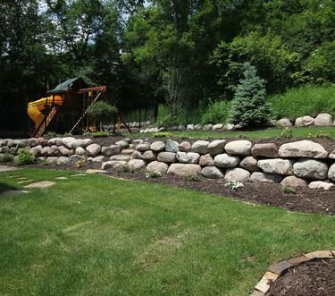 Landscaped backyard with a stone retaining wall, green lawn, trees, and a playground slide in the background