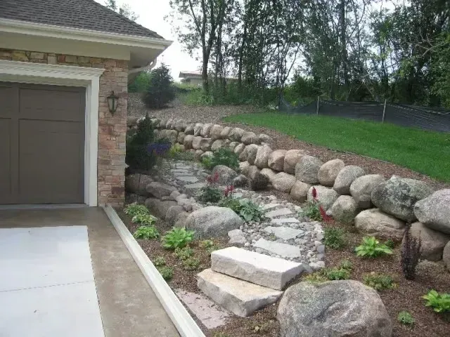 Driveway beside house with stone retaining wall, landscaped slope, and concrete stepping stones