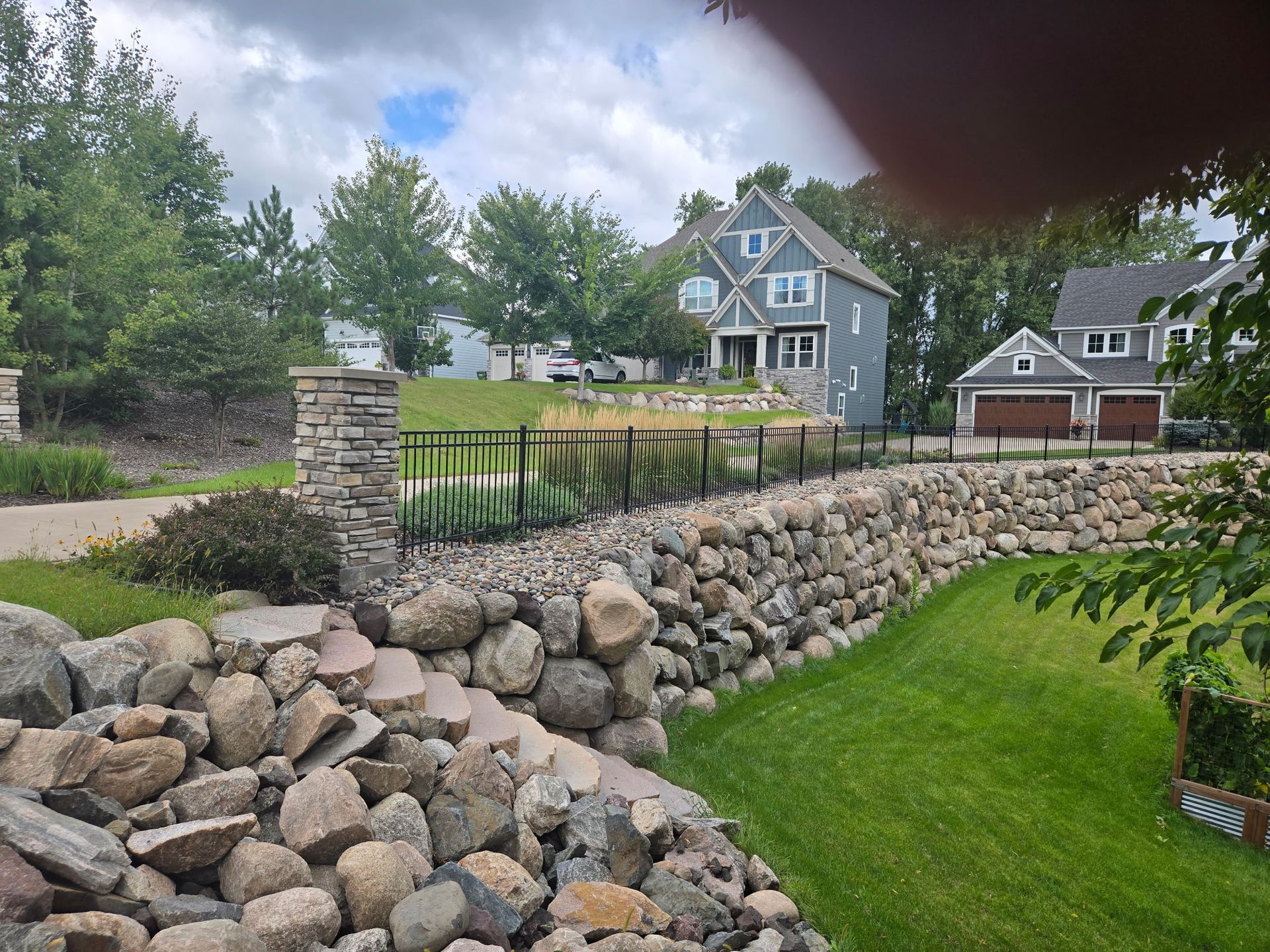 Stone retaining wall and black fence beside a green lawn, with houses and trees in the background.