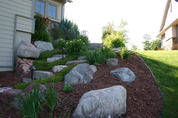 Rock garden with large boulders, mulch, and green shrubs beside a house on a sloped yard