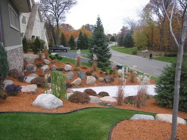 Landscaped front yard with rock garden, shrubs, and a small evergreen beside a suburban road.