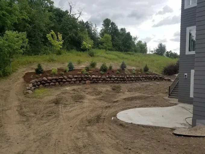 Curved stone retaining wall beside a dirt yard and concrete patio, with trees and a house edge nearby