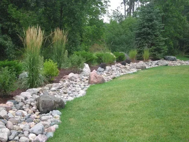 Landscaped garden with a rock border, tall grasses, and a green lawn beside a wooded area