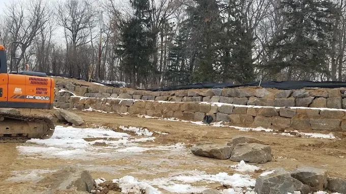 Construction site with stone retaining wall, muddy ground, and orange excavator beside a wooded area.