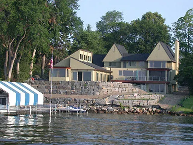 Waterfront lodge with tan buildings, stone retaining wall, and blue-and-white boat cover by calm water