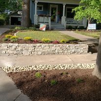Front yard garden with stone retaining wall, concrete steps, and porch of a gray house.