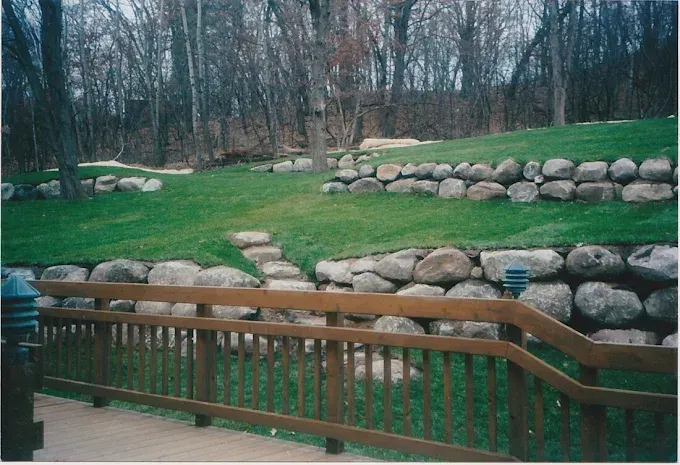 Wooden railing in front of terraced stone retaining walls on a grassy hillside with bare trees.