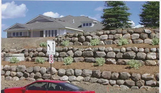 Large house behind stacked stone retaining wall, with a red car partly visible at the bottom.