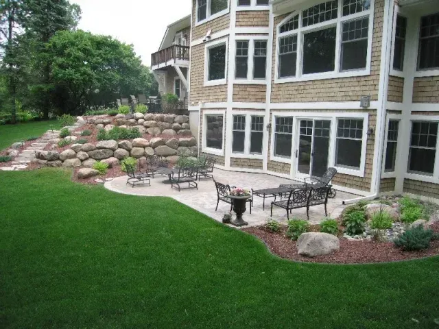 Landscaped backyard patio beside a stone house, with lawn, flowers, and outdoor tables and chairs.