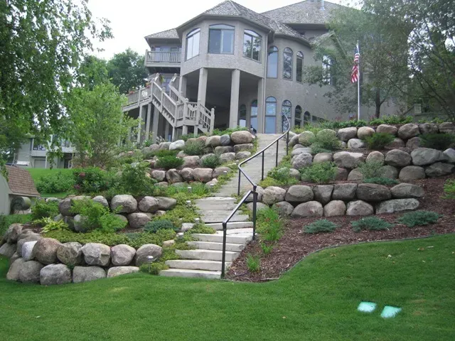Stone steps and terraced rock landscaping leading up to a gray house with a porch and trees.