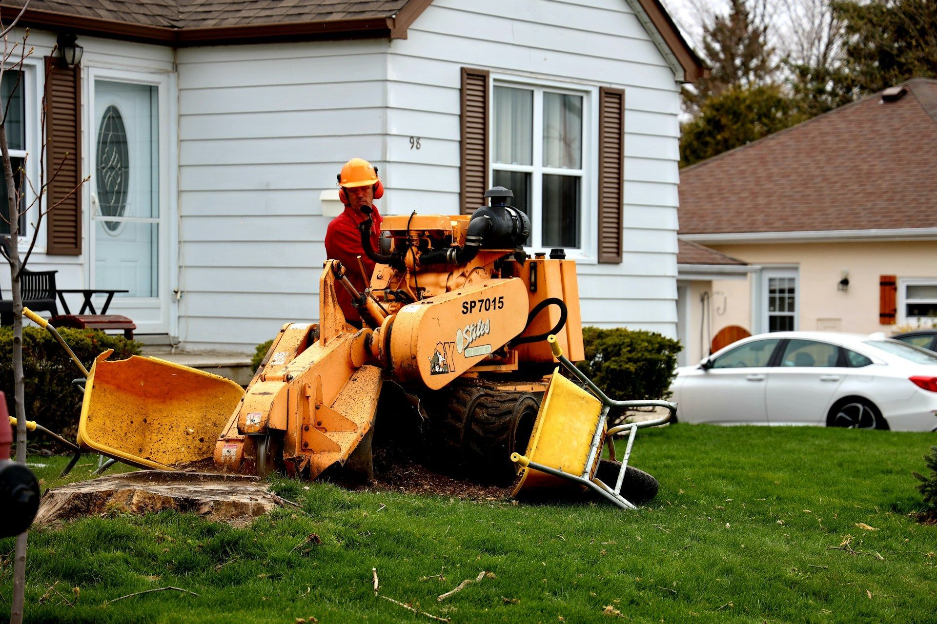 Worker operating a yellow snow blower near a white house with a green lawn.