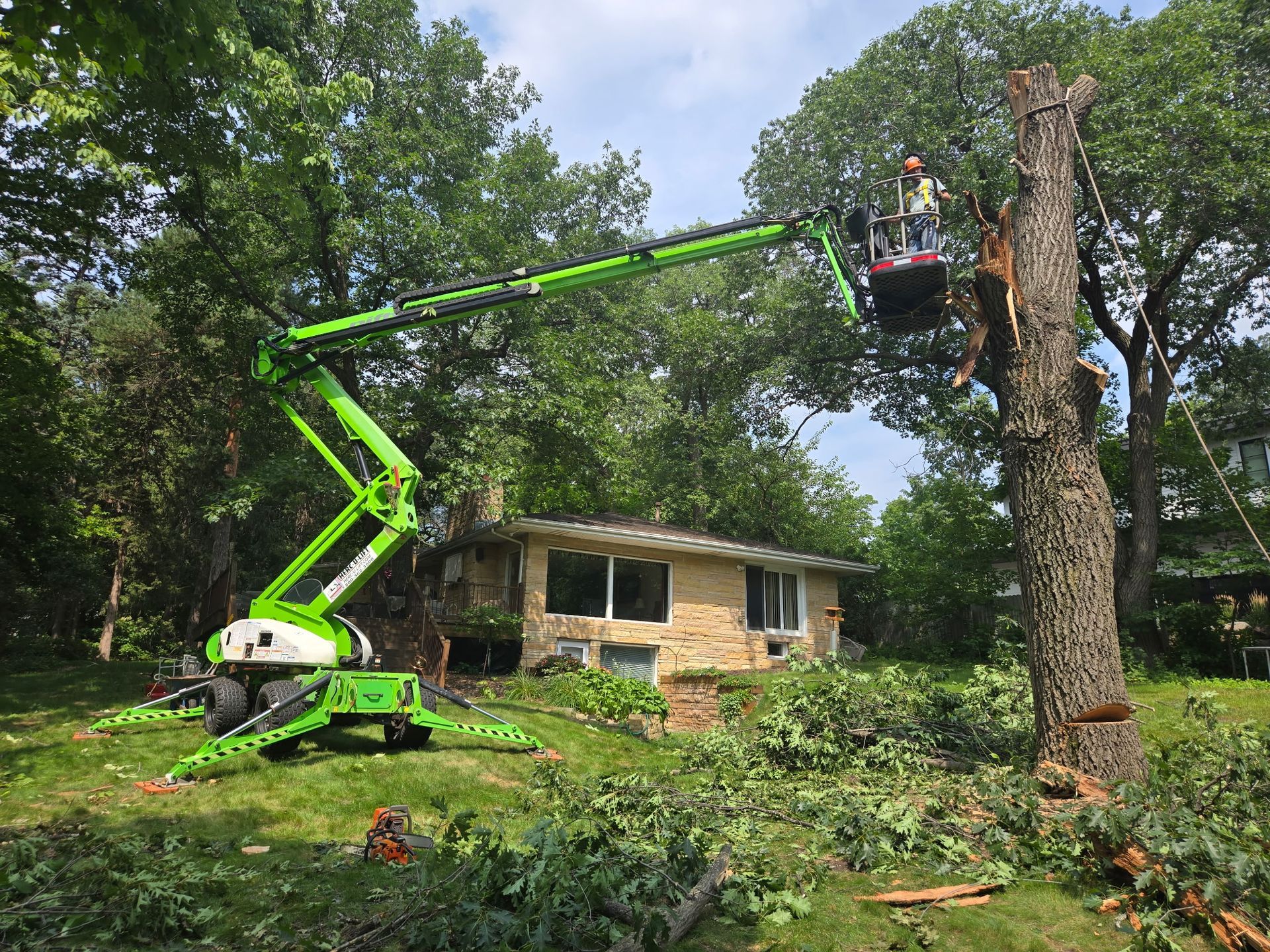 Green bucket truck trims a large tree beside a small house after storm damage.