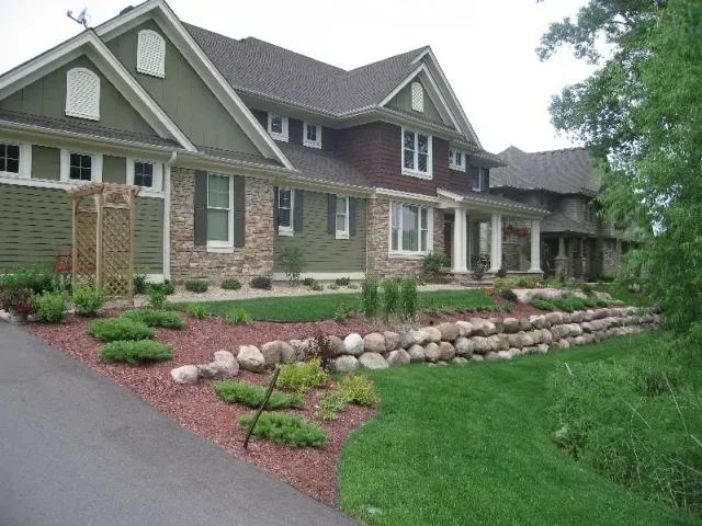 Large suburban house with stone and green siding, landscaped front yard, and a sloped driveway