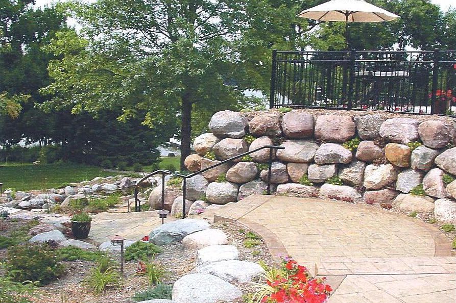 Stone patio with stacked rock wall, black railing, and beige umbrella beside a landscaped garden