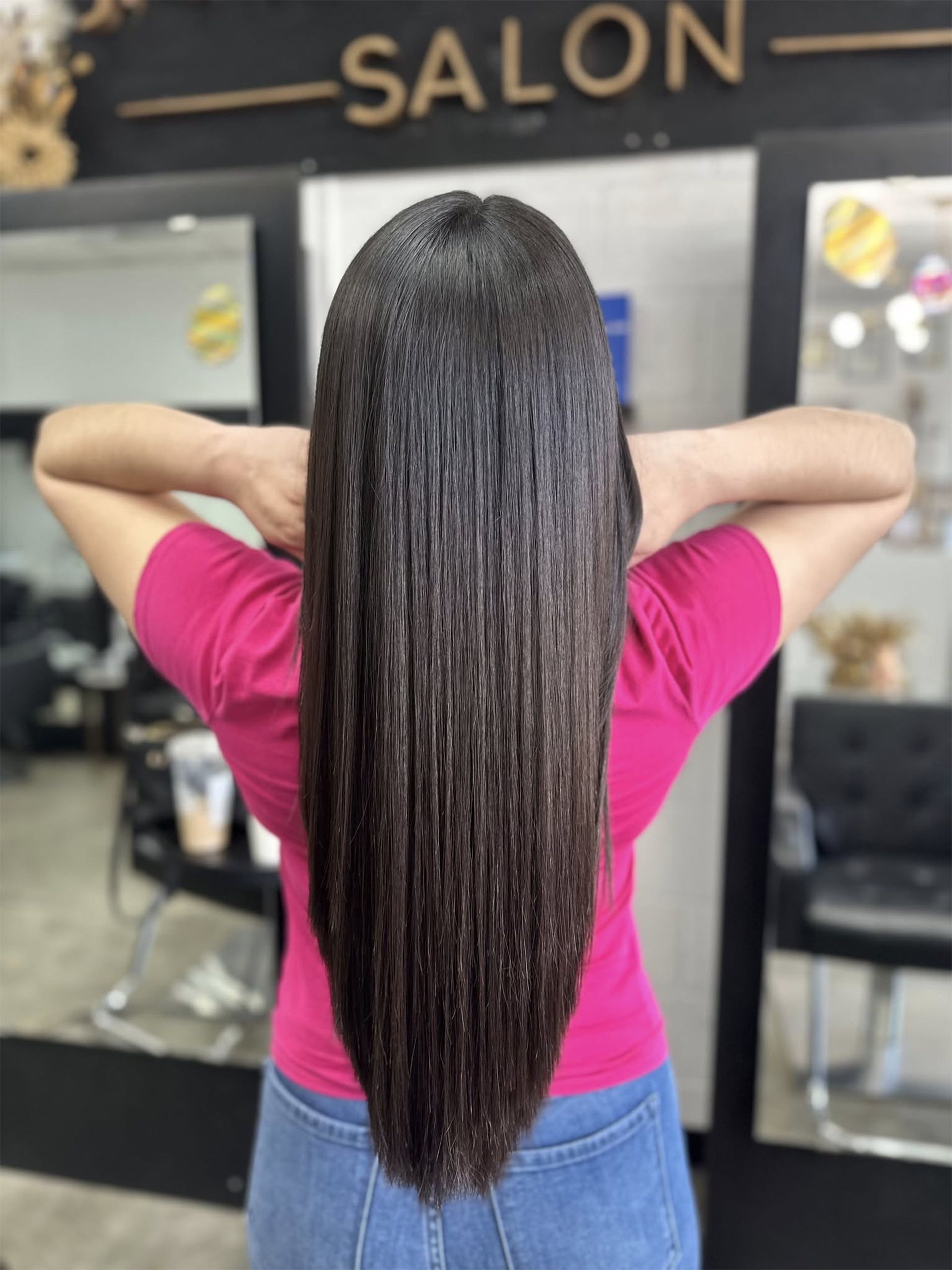 Woman with long, sleek, dark hair after a salon visit. She wears a pink shirt and stands in front of a salon.