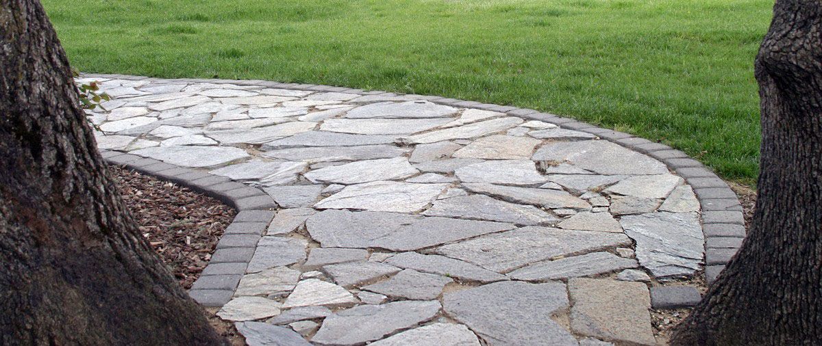 A stone walkway leading to a lush green field surrounded by trees.