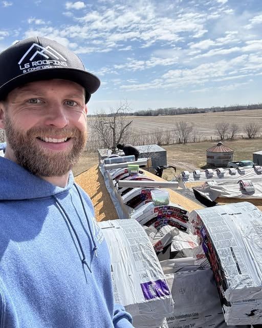 Smiling man in a blue hoodie and cap standing by stacked roof shingles on a rooftop with fields in the background