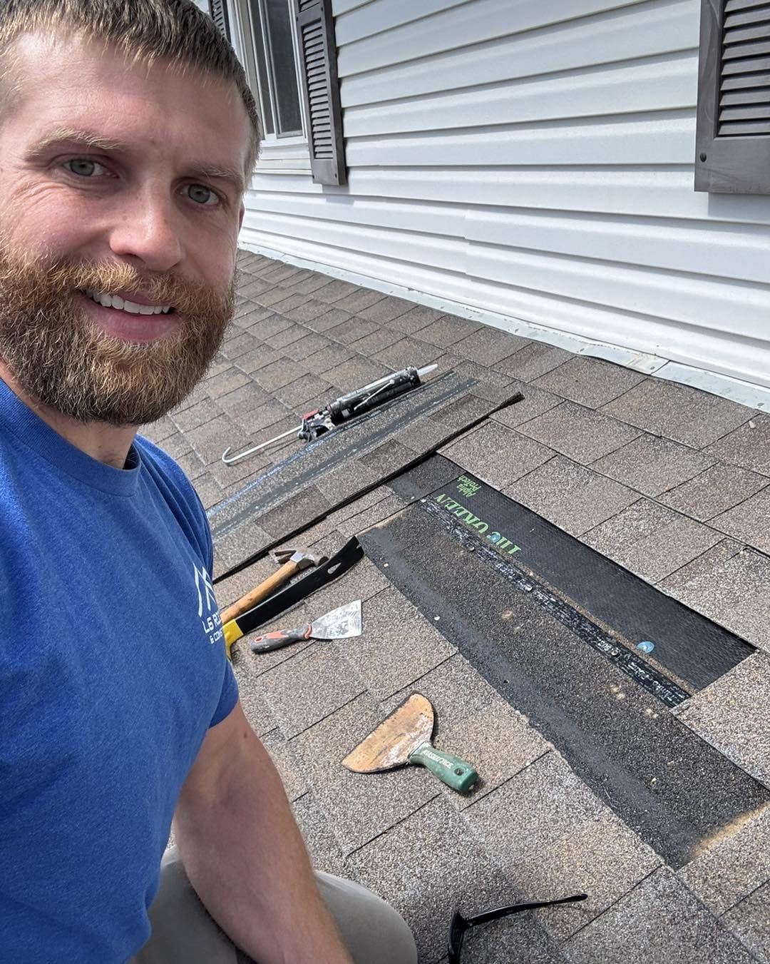 Man in blue shirt on shingled roof beside roofing tools and a patch of black underlayment