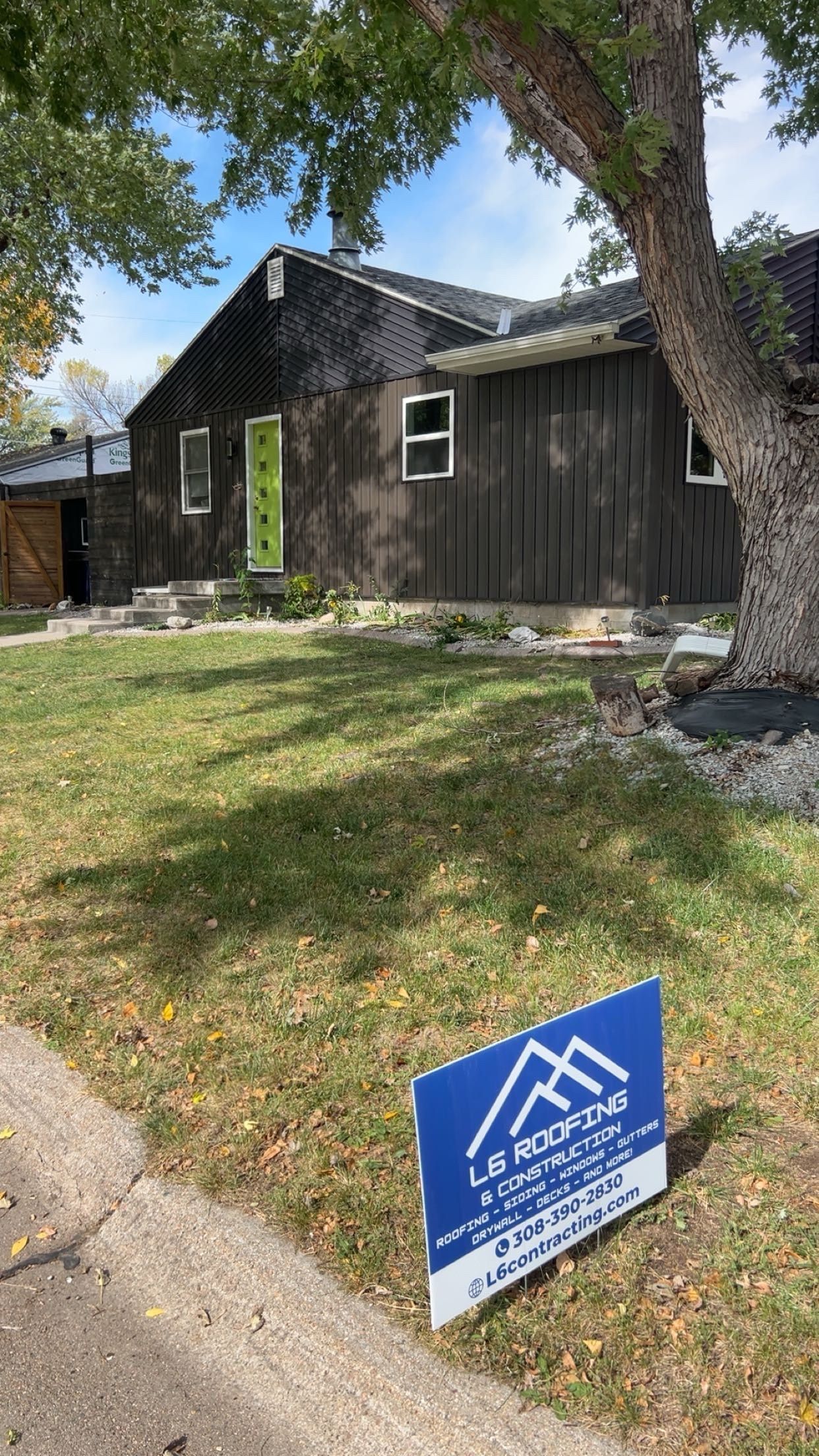 Small dark house with green door behind a blue yard sign under trees in a grassy front yard.