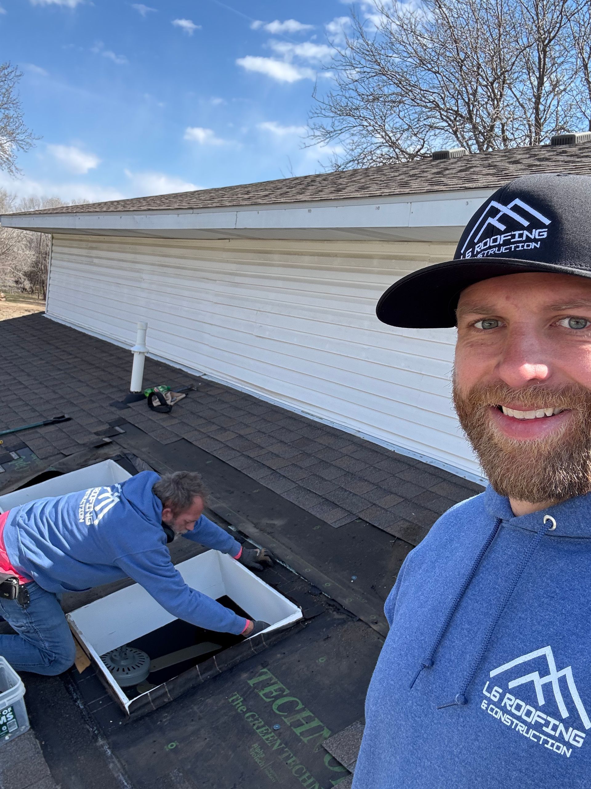 Two workers inspecting a flat roof, one smiling selfie-style in foreground under a blue sky.