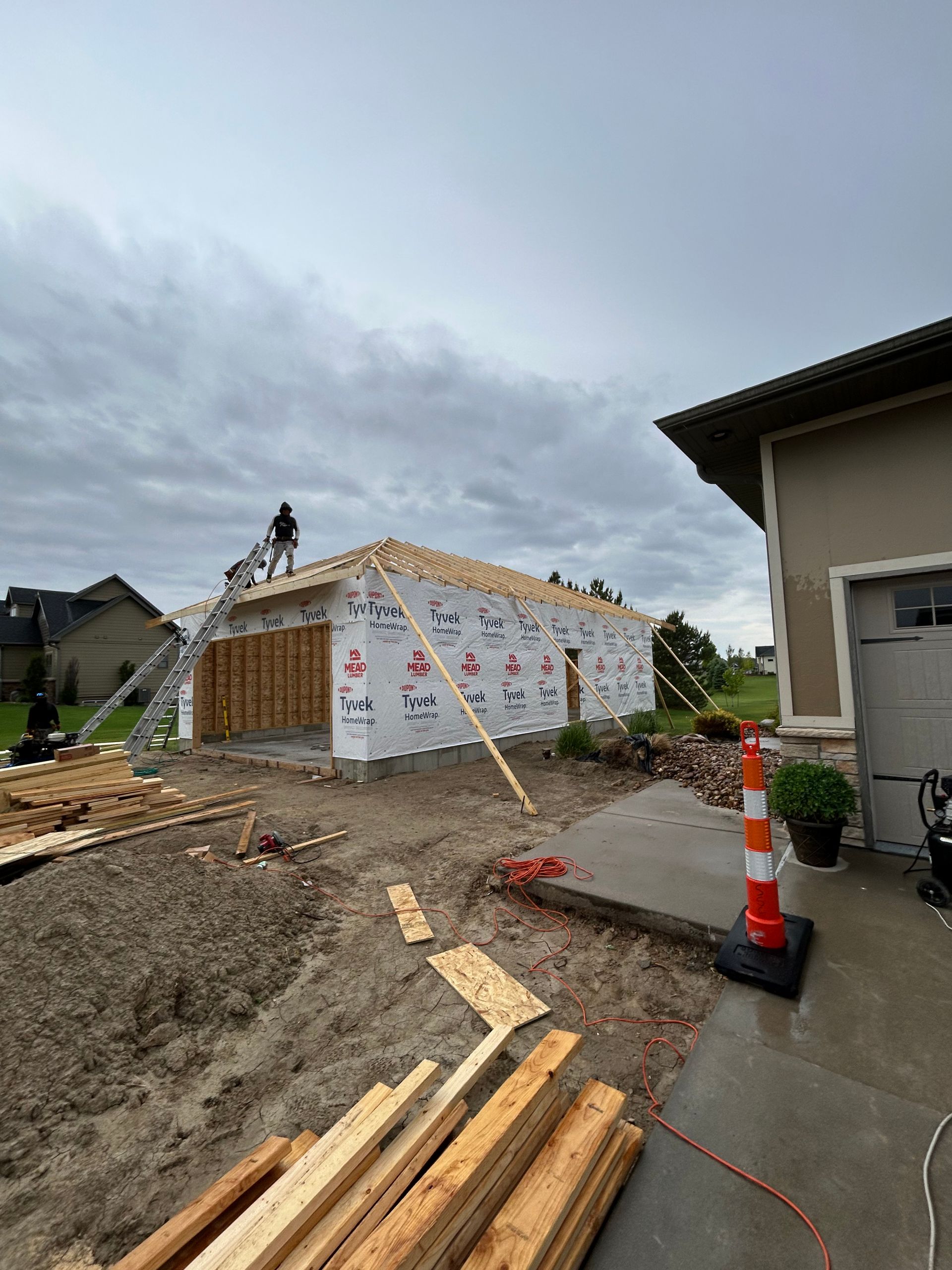 House under construction with roof framing, lumber, and an orange traffic cone on a muddy site