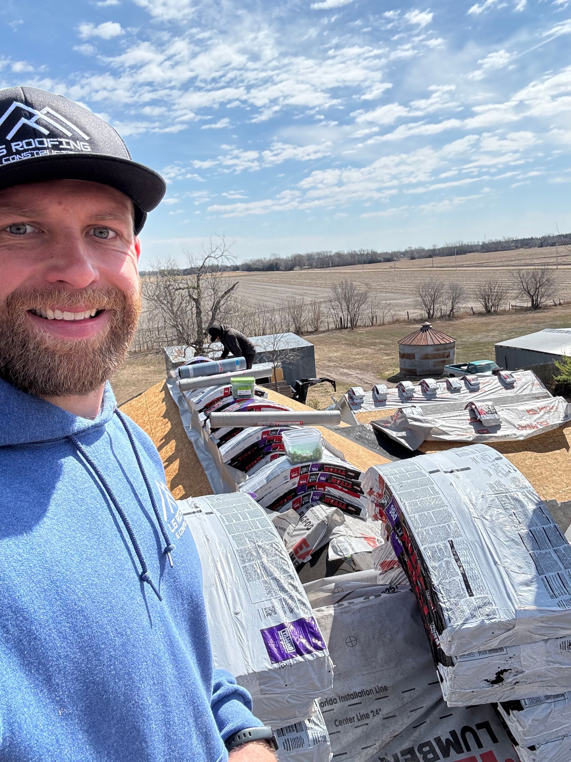 Selfie of a man in a blue shirt beside stacks of bagged materials in a snowy field under a cloudy sky
