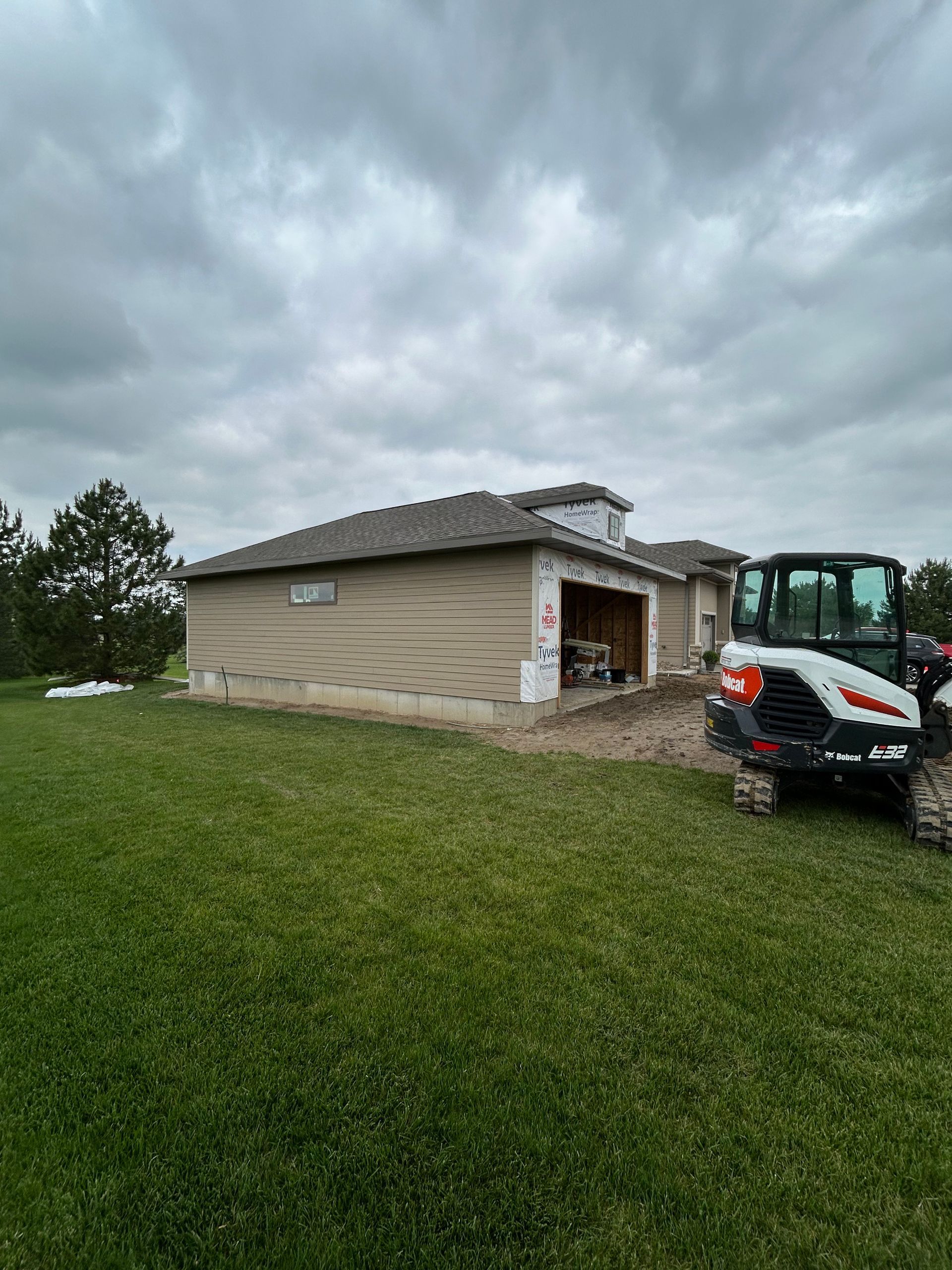Small beige house with a Bobcat machine on a grassy yard under cloudy skies.