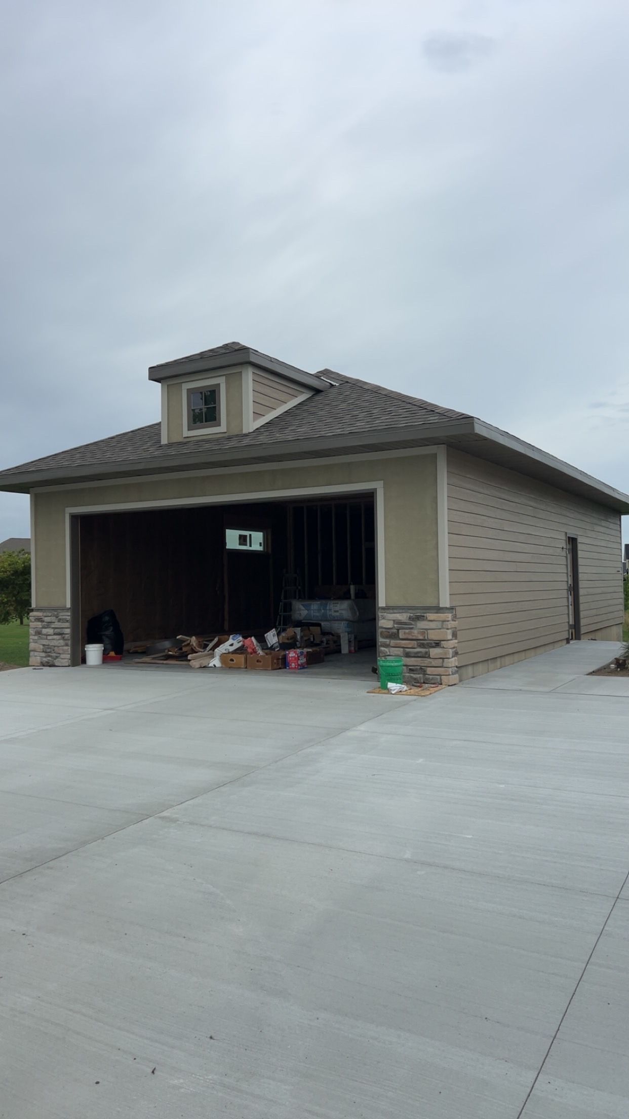 Suburban garage with open door, clutter inside, and a large concrete driveway under cloudy skies