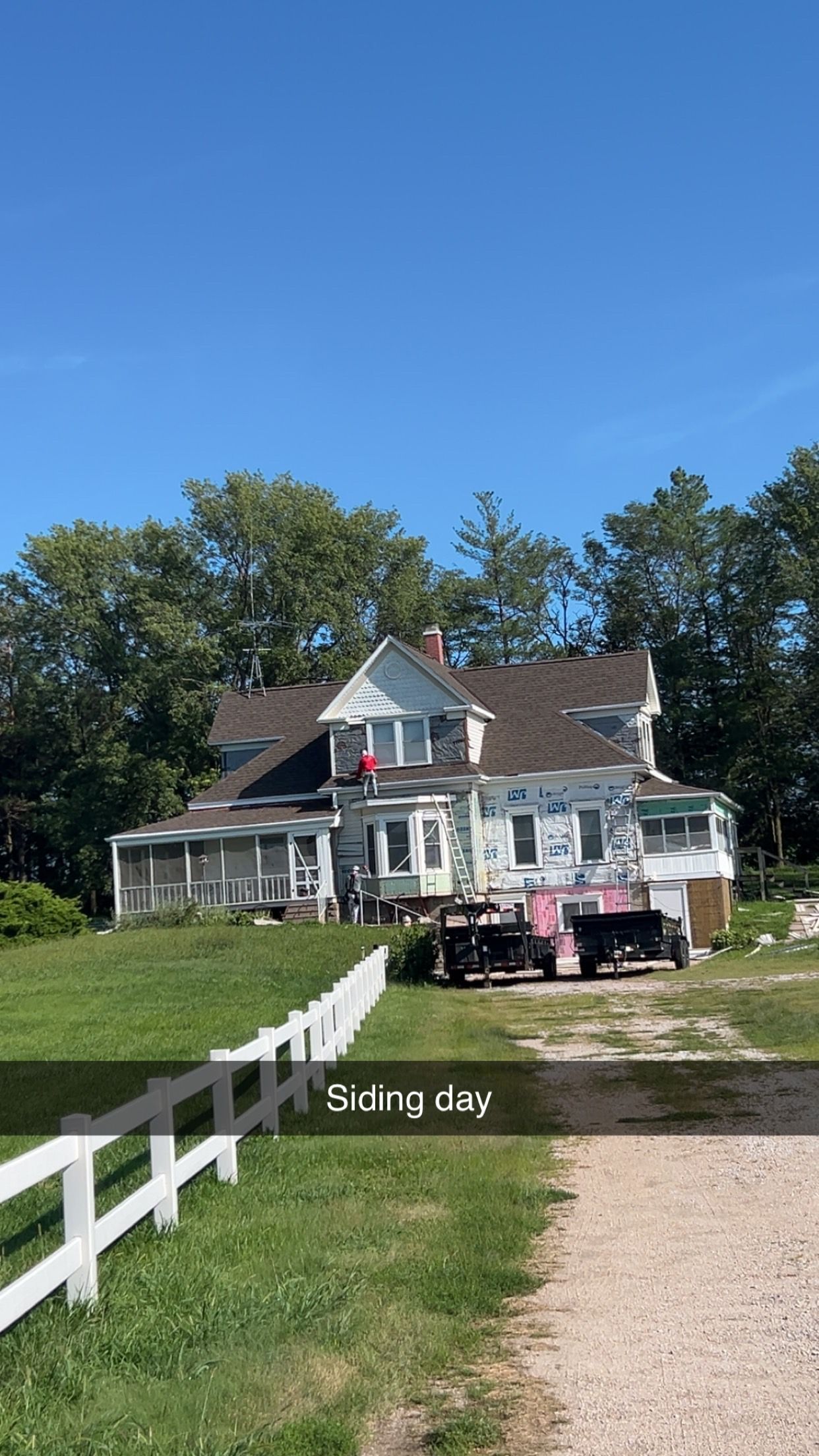 Old white farmhouse with red trim beside a gravel driveway under a clear blue sky.