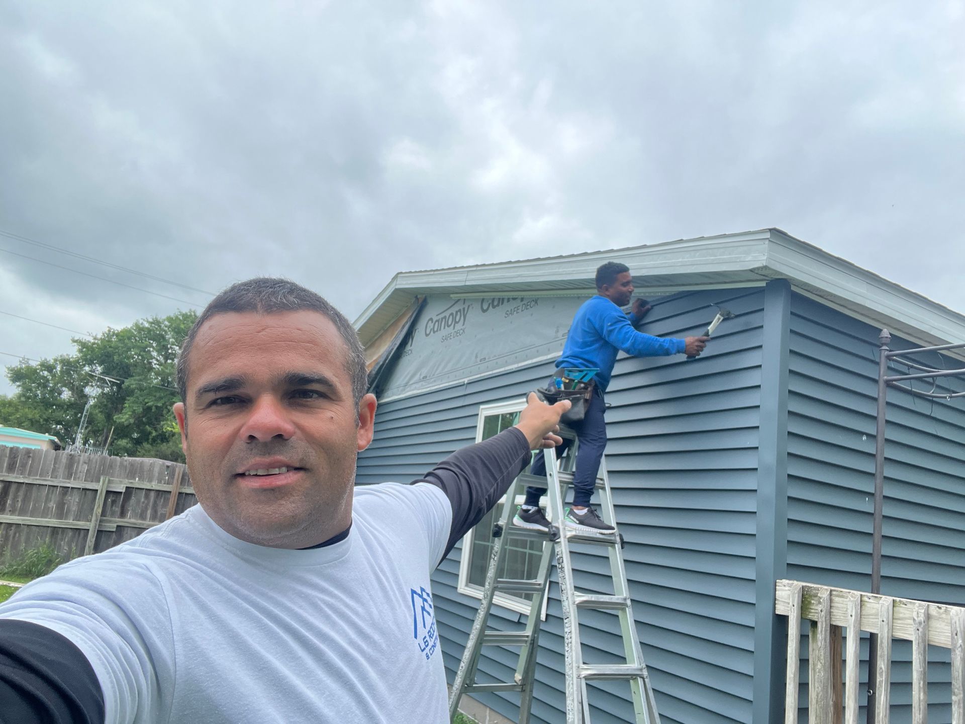 Man taking a selfie while a worker paints the side of a gray house on a ladder