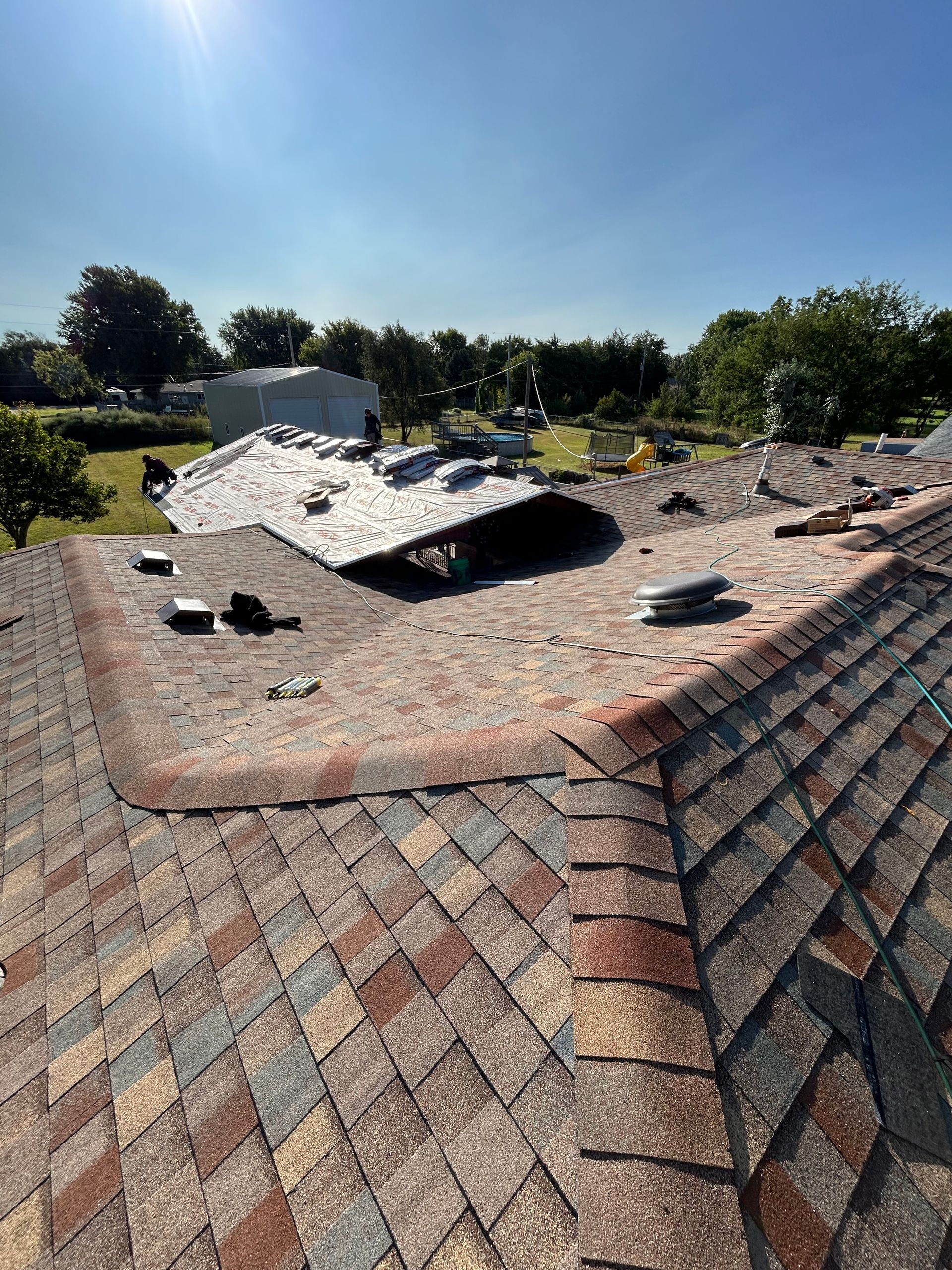 Damaged shingle roof with a large metal vent or cap lifted open under a clear blue sky
