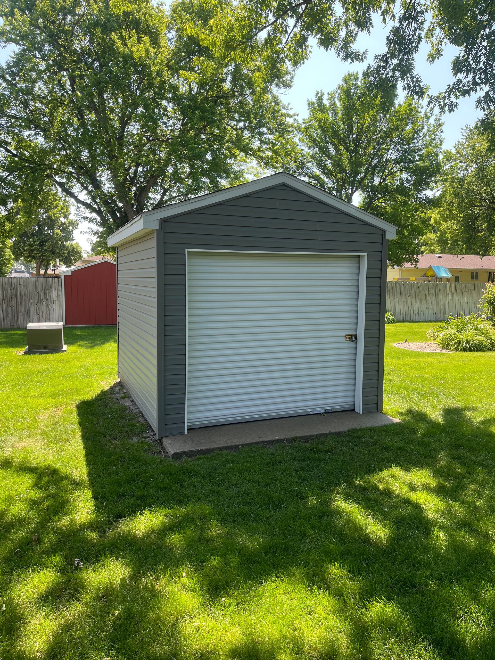 Small gray shed with a white roll-up door in a grassy backyard under trees.