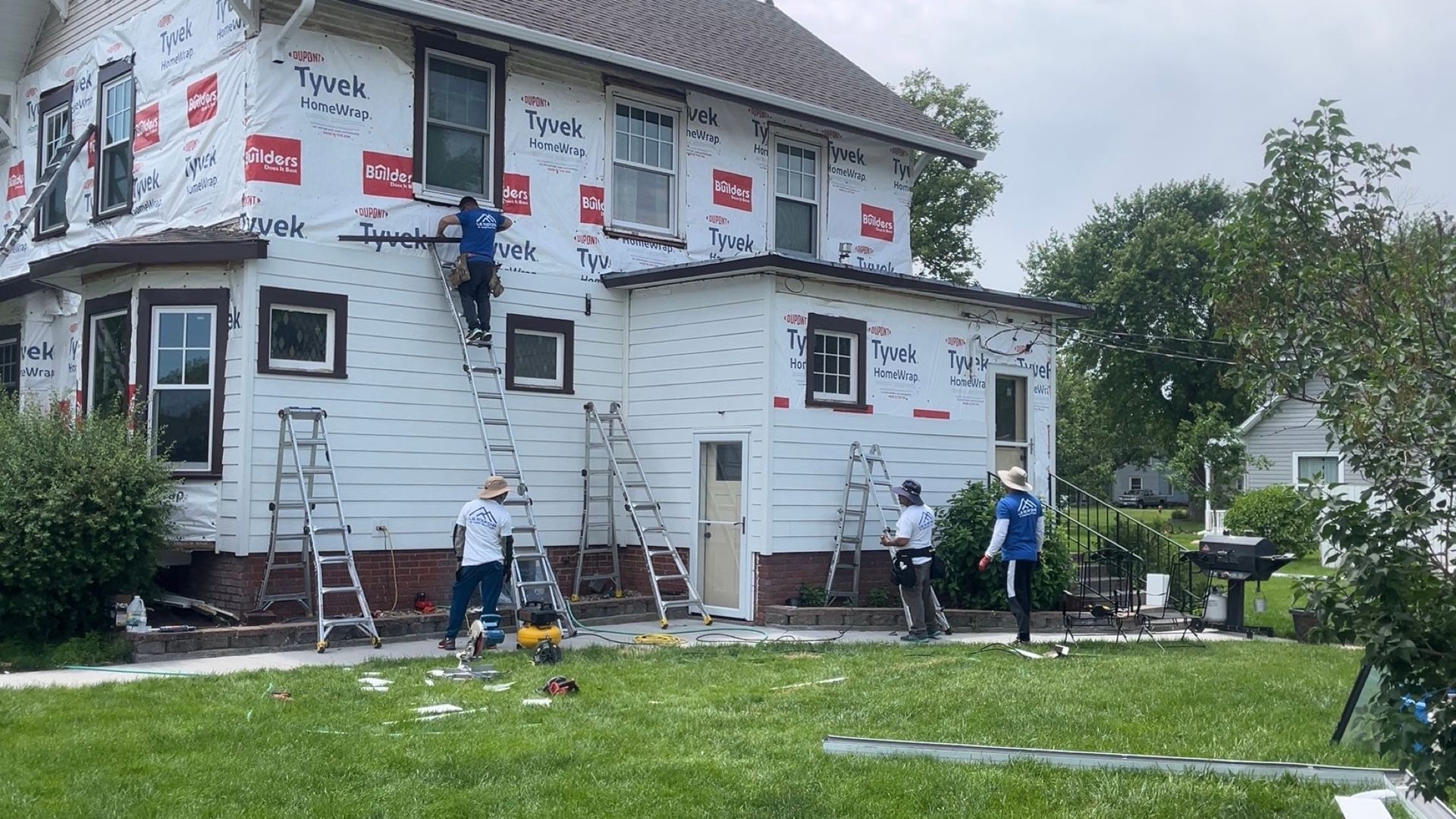 Workers on ladders insulating a white house exterior, with construction materials around the yard.