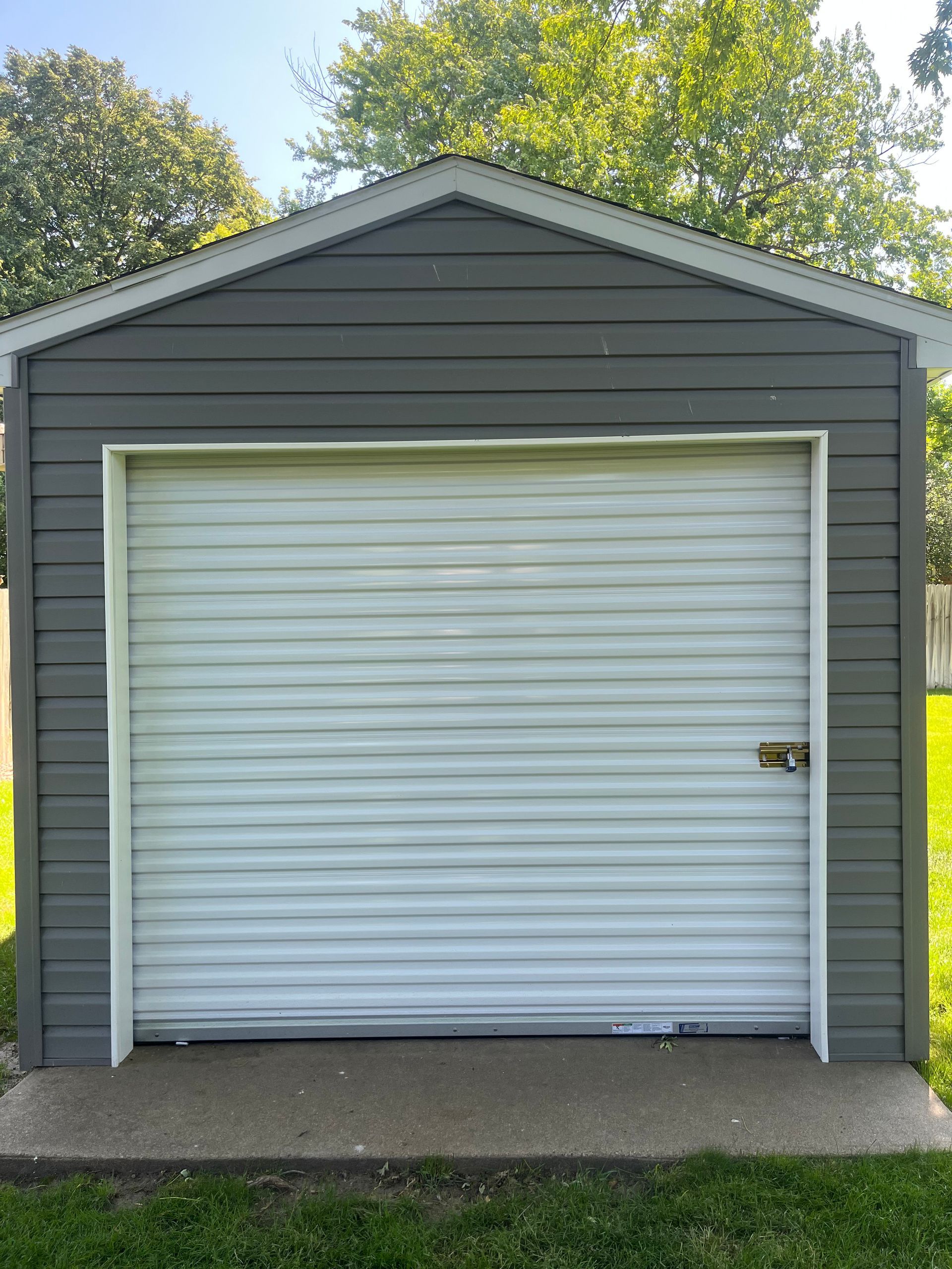 Gray garage with white roll-up door, centered on a concrete slab with trees in the background