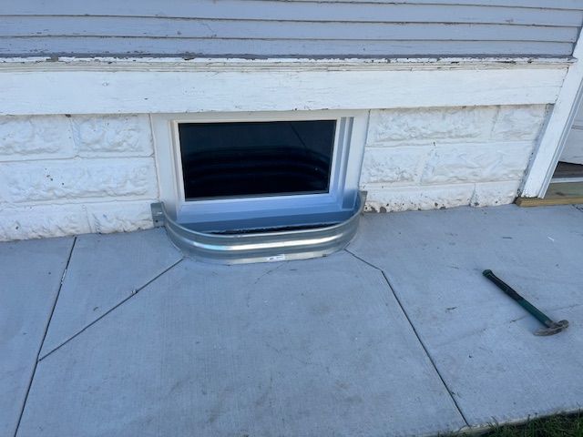 Basement window with metal well cover beneath house siding, on concrete patio.