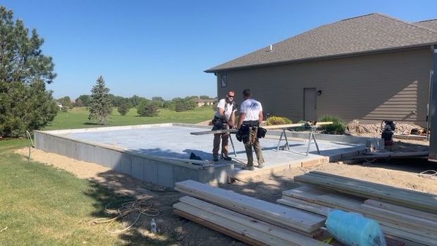 Workers pouring concrete patio beside a house, with tools and lumber in the foreground.