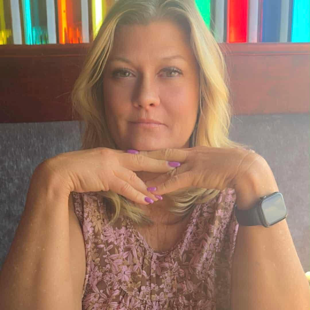 Woman with blonde hair, resting chin on hands, wearing a floral shirt and watch. Background has colorful glass.