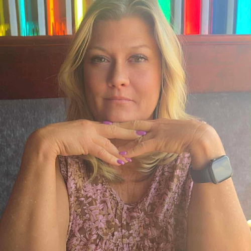 Woman with blonde hair, resting chin on hands, wearing a floral shirt and watch. Background has colorful glass.