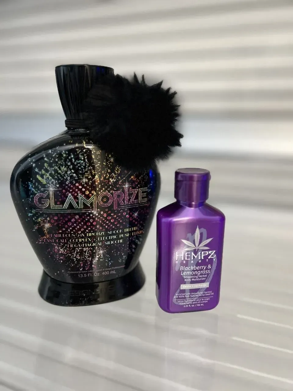 Two tanning lotion bottles: Glamour and Hempz. Glamour is black with a puff. Hempz is purple.