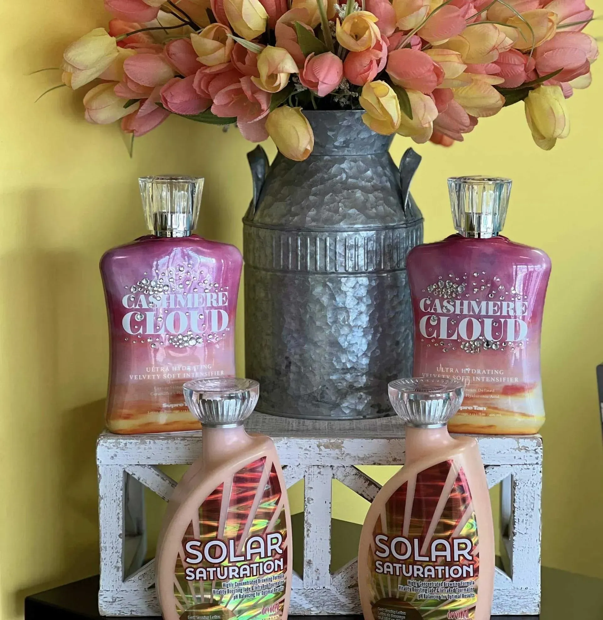 Two bottles of Cashmere Cloud and Solar Sensation lotion arranged with a metal vase and pink and yellow flowers.