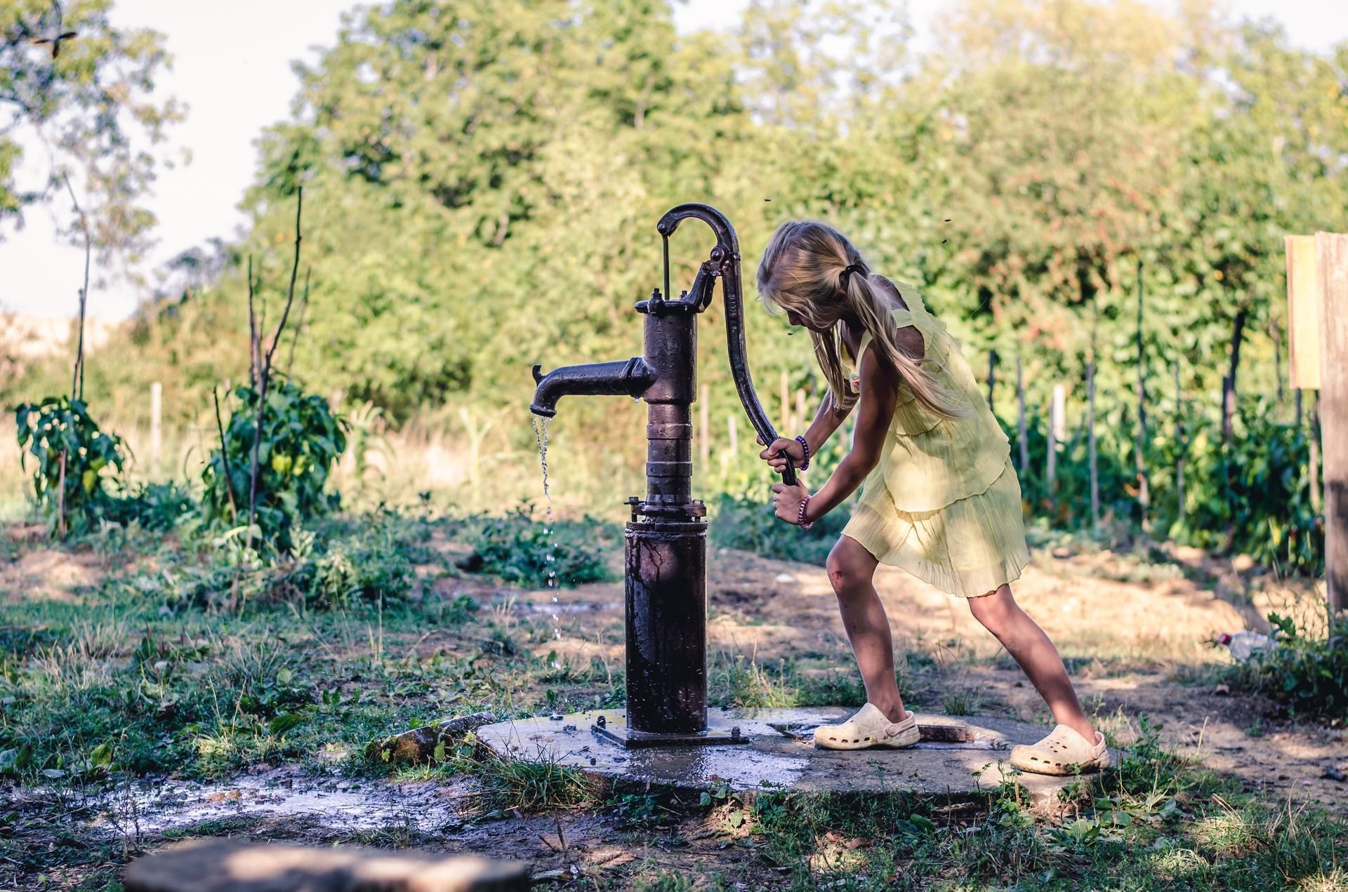 A person in a yellow dress pumps water from an old, rusted metal pump in a sunny garden.