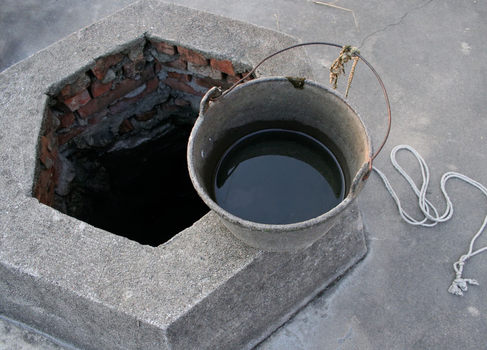 A grey concrete well opening next to a metal bucket filled with water and a length of white rope.